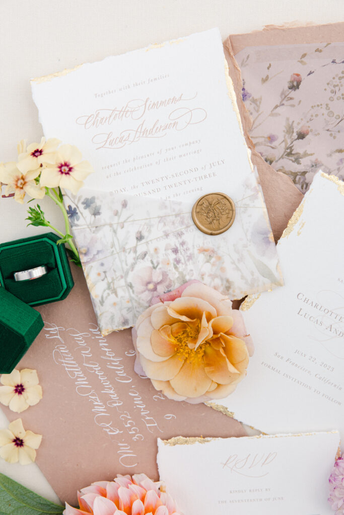 Flatlay with custom wedding invitation with coral and lavender floral designs. Blush handmade paper envelopes with white handwritten calligraphy.