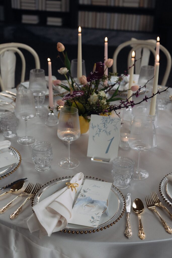 Bow themed wedding table decor with blue illustrated table number and menus with place cards on top of gold plates.