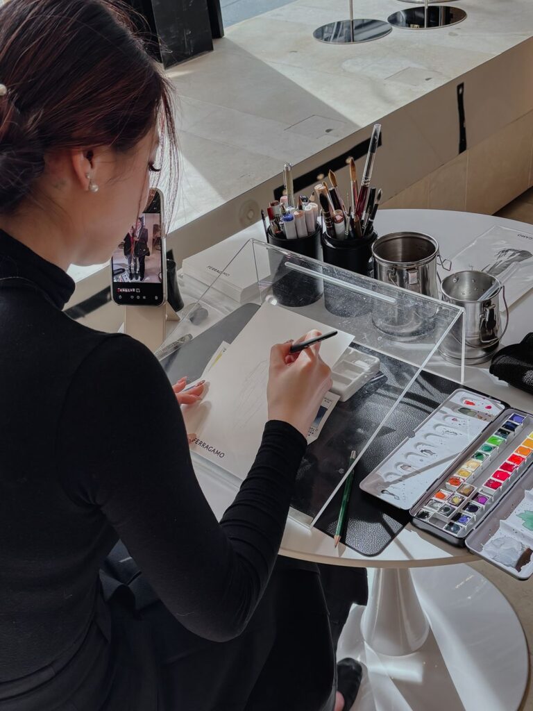 A woman sitting at a table fashion sketching with art supplies surrounding her.